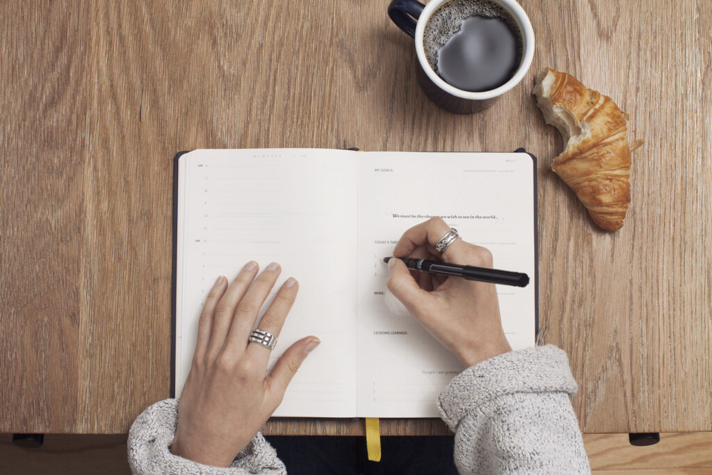 Woman Writing in Notebook with Coffee and Croissant on Desk