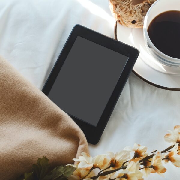 An eReader, next to a blanket, some flowers, and cup of tea and a muffin