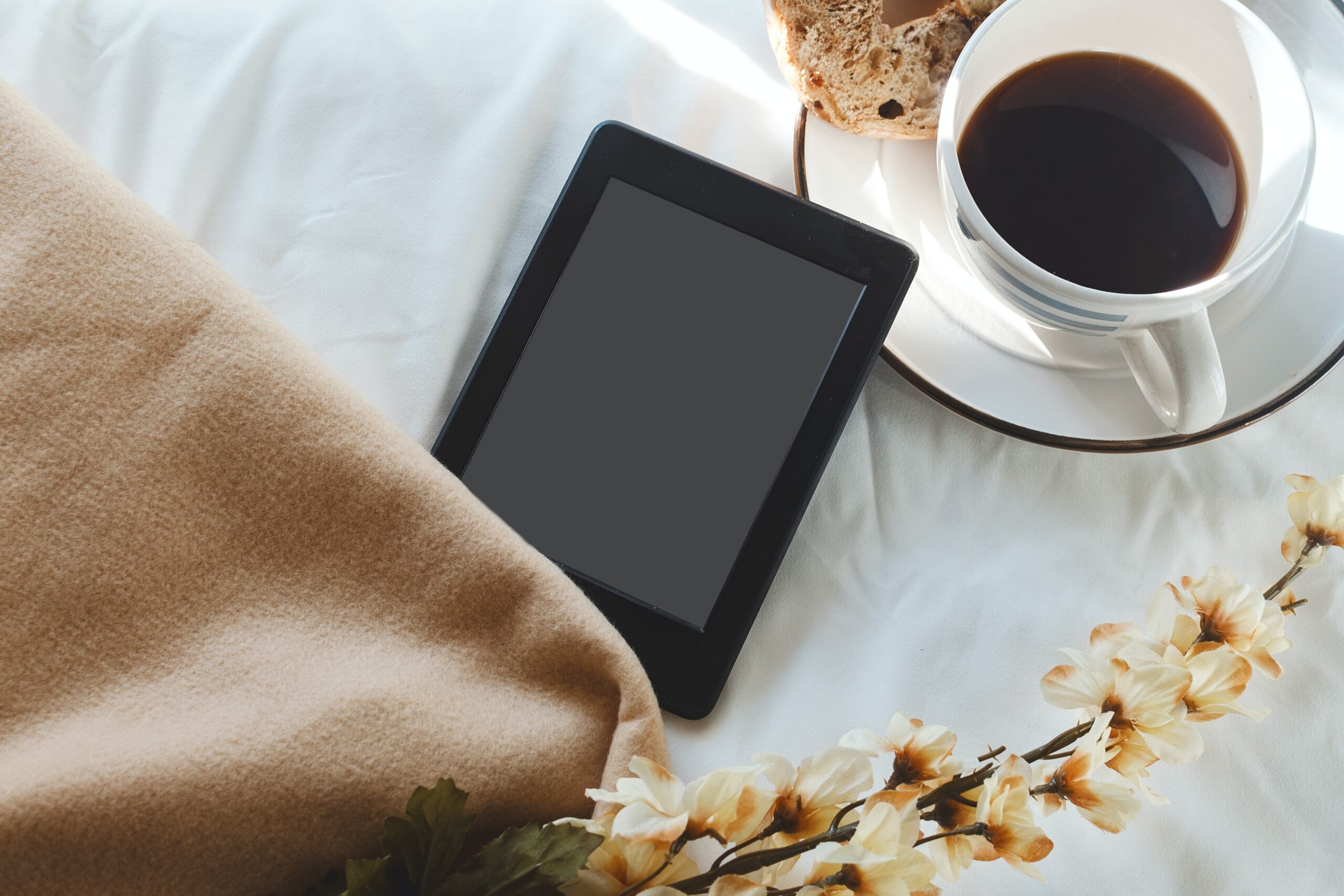 An eReader, next to a blanket, some flowers, and cup of tea and a muffin