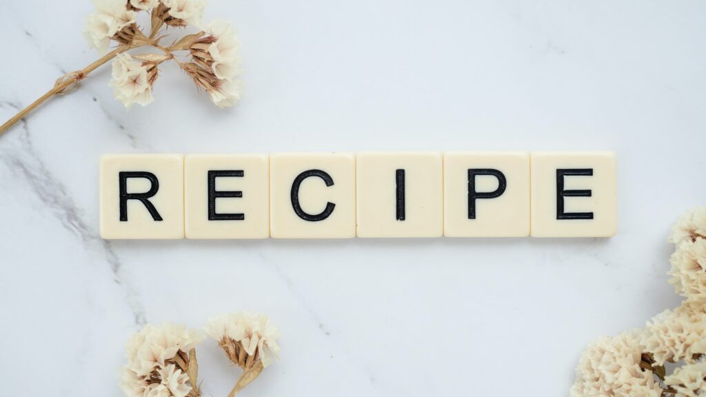 Scrabble pieces spelling out "recipe" with a marble background, surrounded by pale flowers