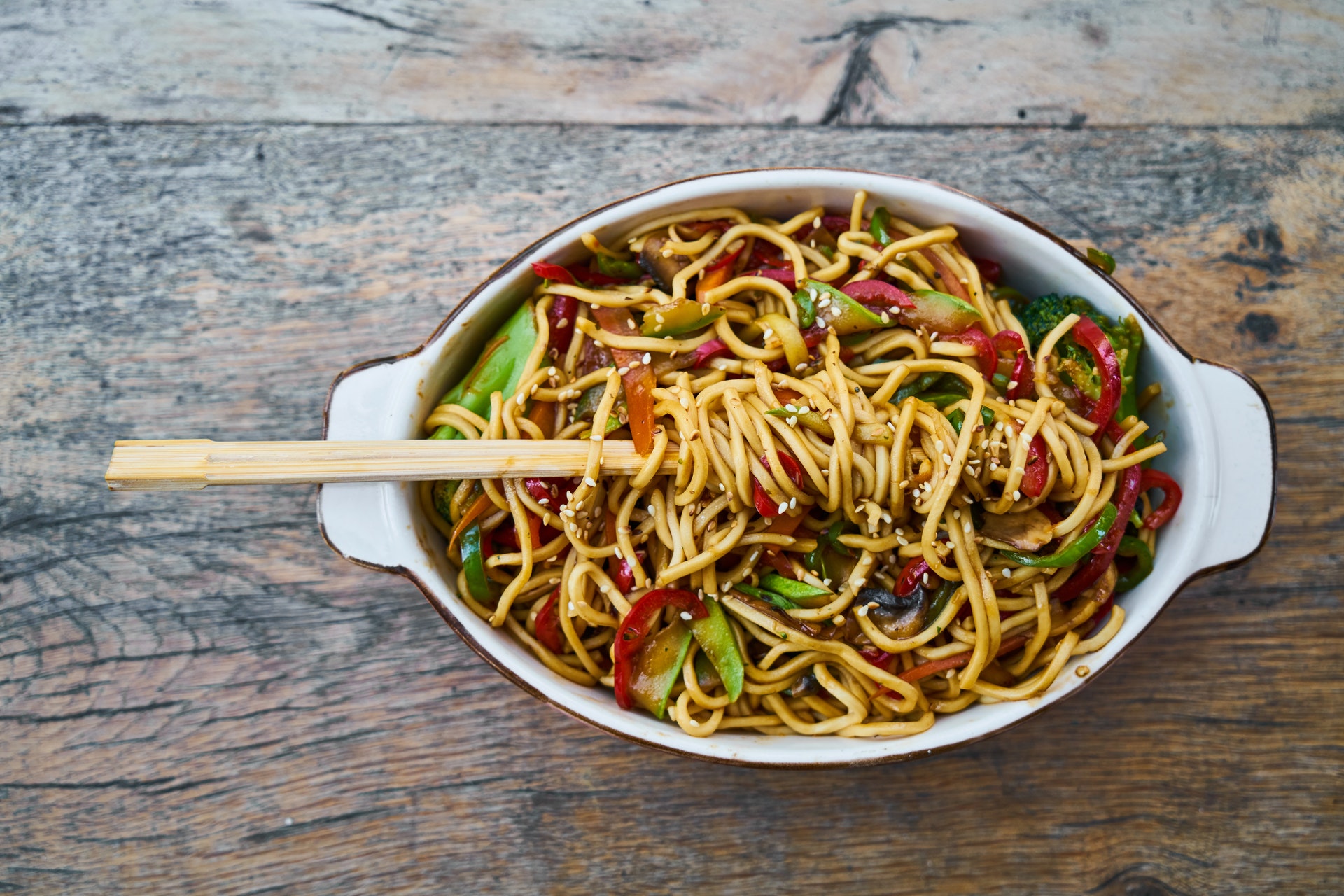 A bowl of stir fried noodles and vegetables with chopsticks on a wooden table