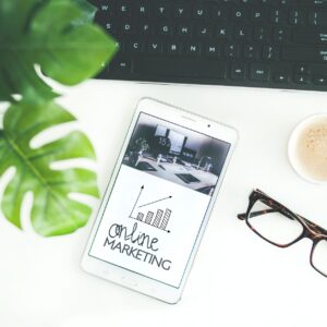 Photo of a laptop, glasses, cup of coffe, a plant, and phone with the words online marketing on the screen