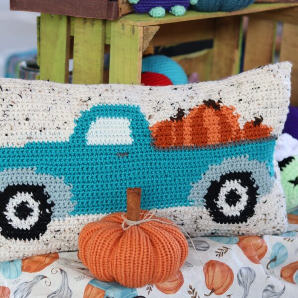 A throw pillow with a blue pickup truck and orange pumpkins crocheted on it