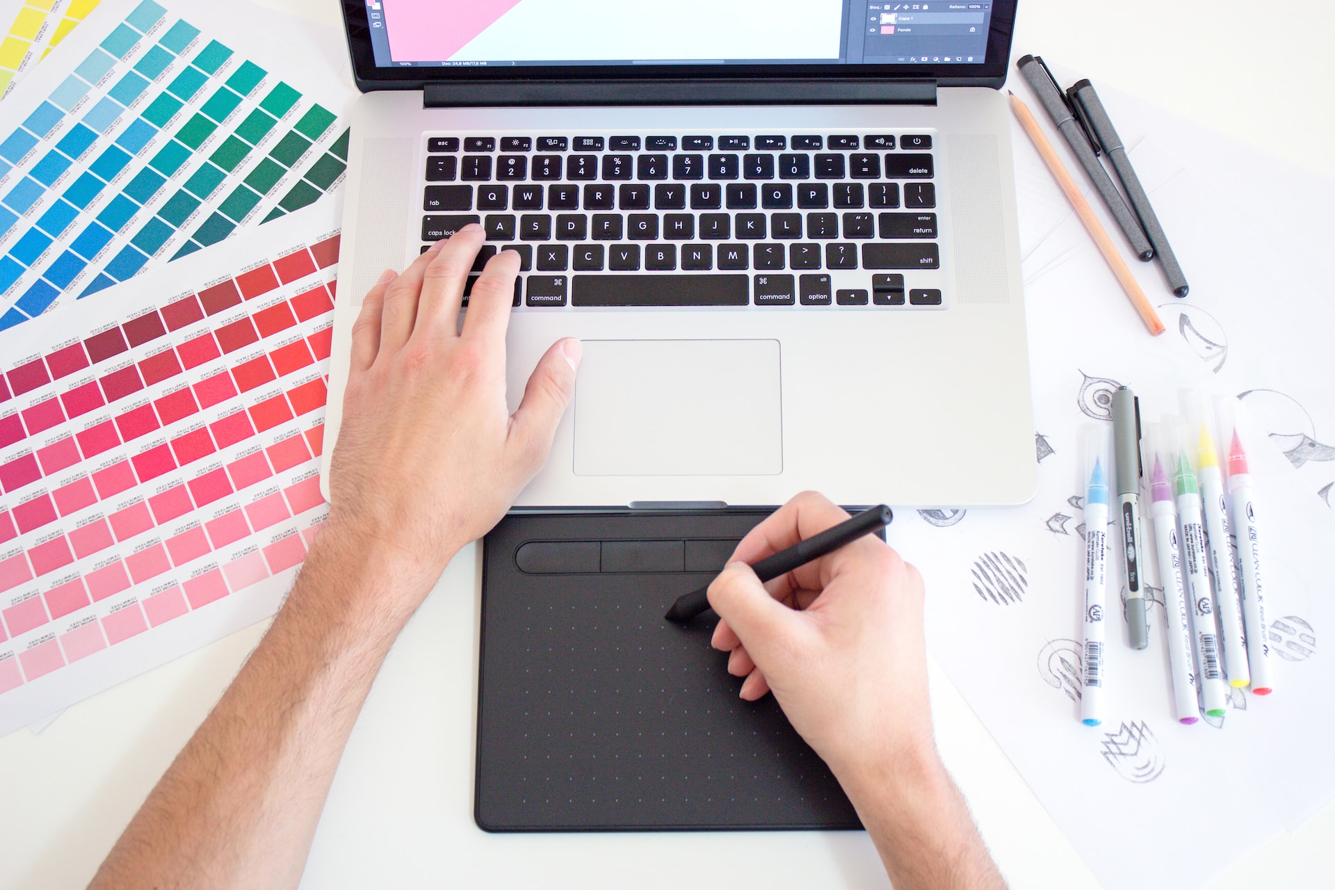 Graphic designer working on a MacBook laptop using a trackpad, colour charts and markers