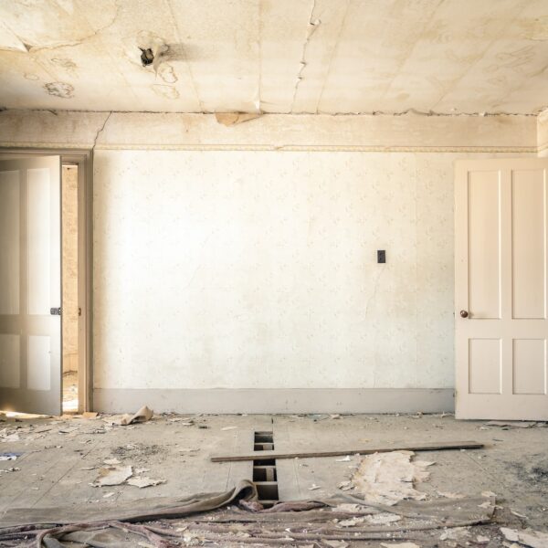 Room with two doors, stained wallpaper, damp ceiling, and a floorboard that has been removed