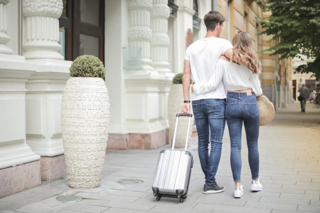 A man and a woman both wearing white shirts walking on the pavement and wheeling a suitcase