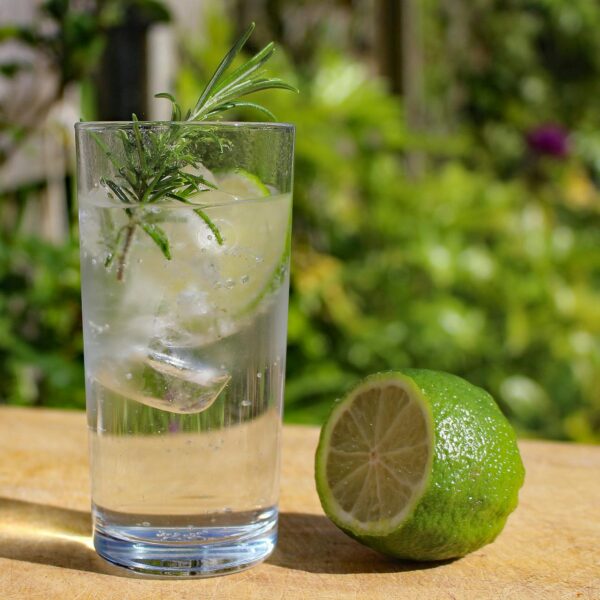A glass of gin and tonic with ice and a sprig of rosemary next to a lime on a wooden table outdoors